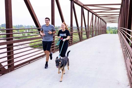 Man and woman crossing bridge over Boise river with dog on leash.