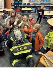 Firefighter kneeling down with small children feeling his helmet. 