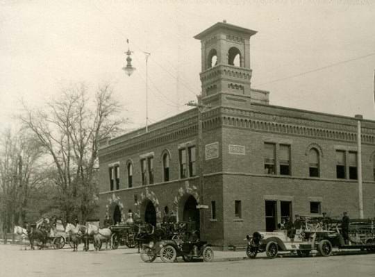 Photo from around 1912. 2 story brick building with bell tower featuring horse-pulled fire wagons.