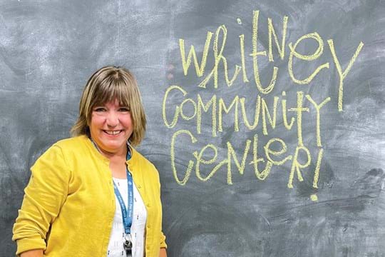 Smiling woman in a yellow sweater stands in front of a blackboard that says Whitney Community Center