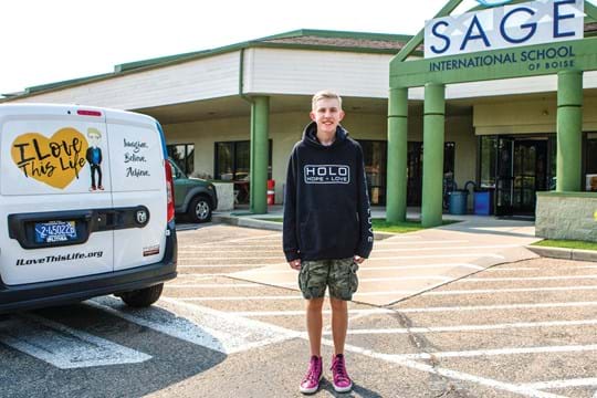 Teenage boy stands in front of a van and a high school.