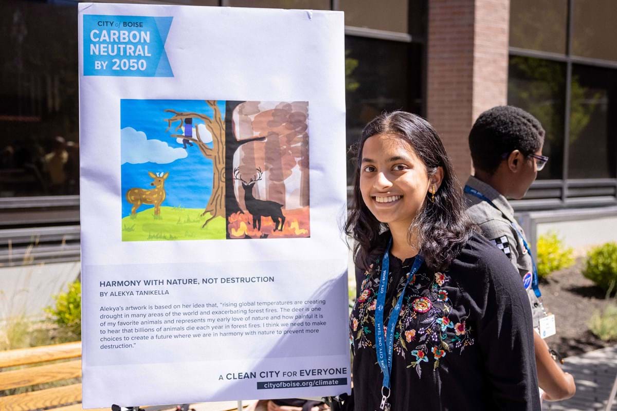 Girl standing next to her artwork depicting a deer and a tree before and after a forest fire.