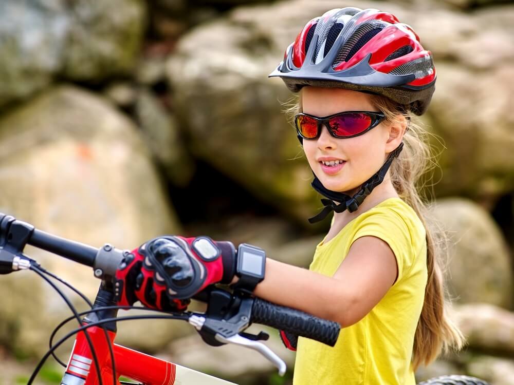 A young girl wearing yellow shirt with helmet and mountain biking gloves, holding a mountain bike up.