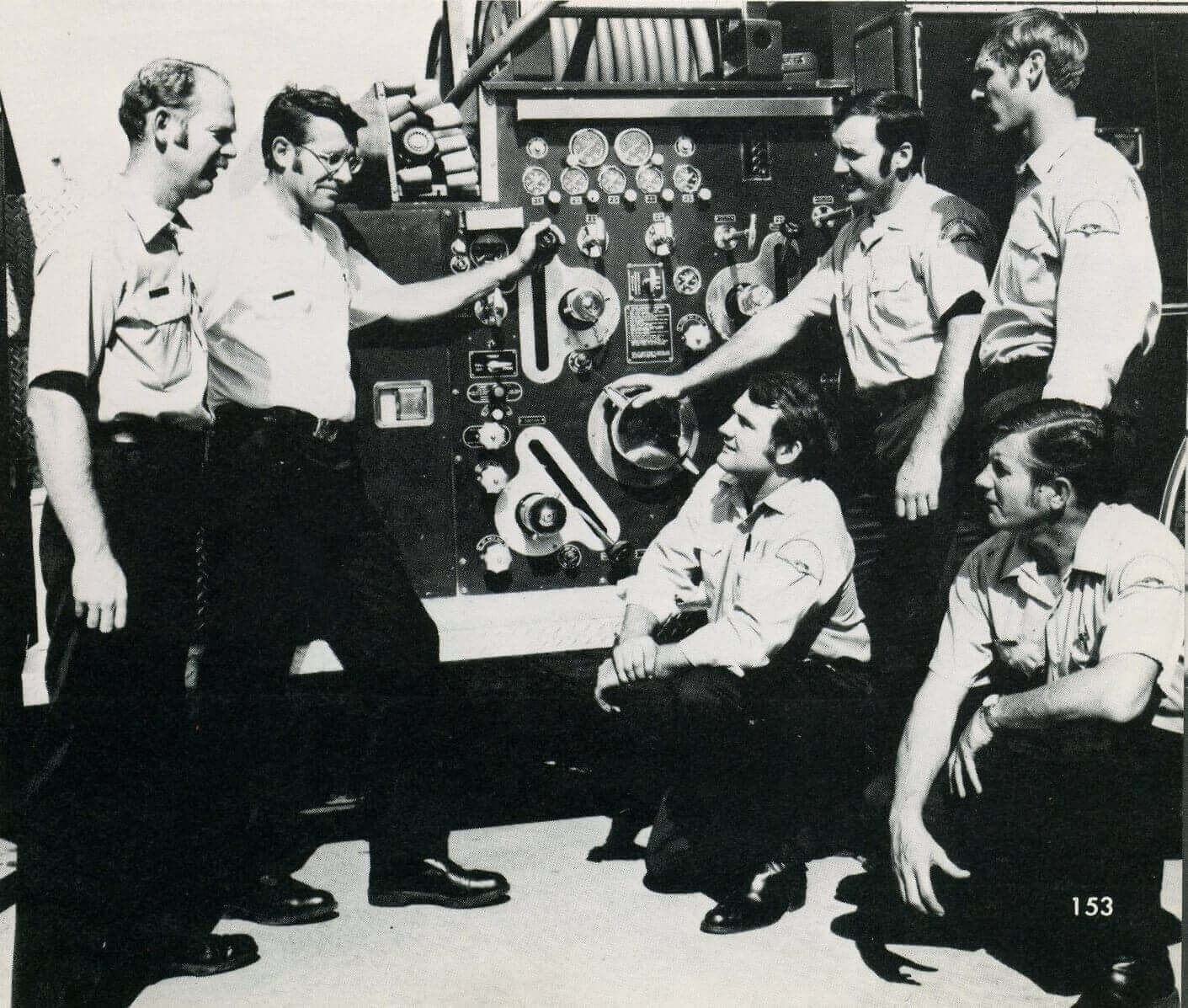 7 firefighters around the pump area of a truck - photo taken in the 1970s
