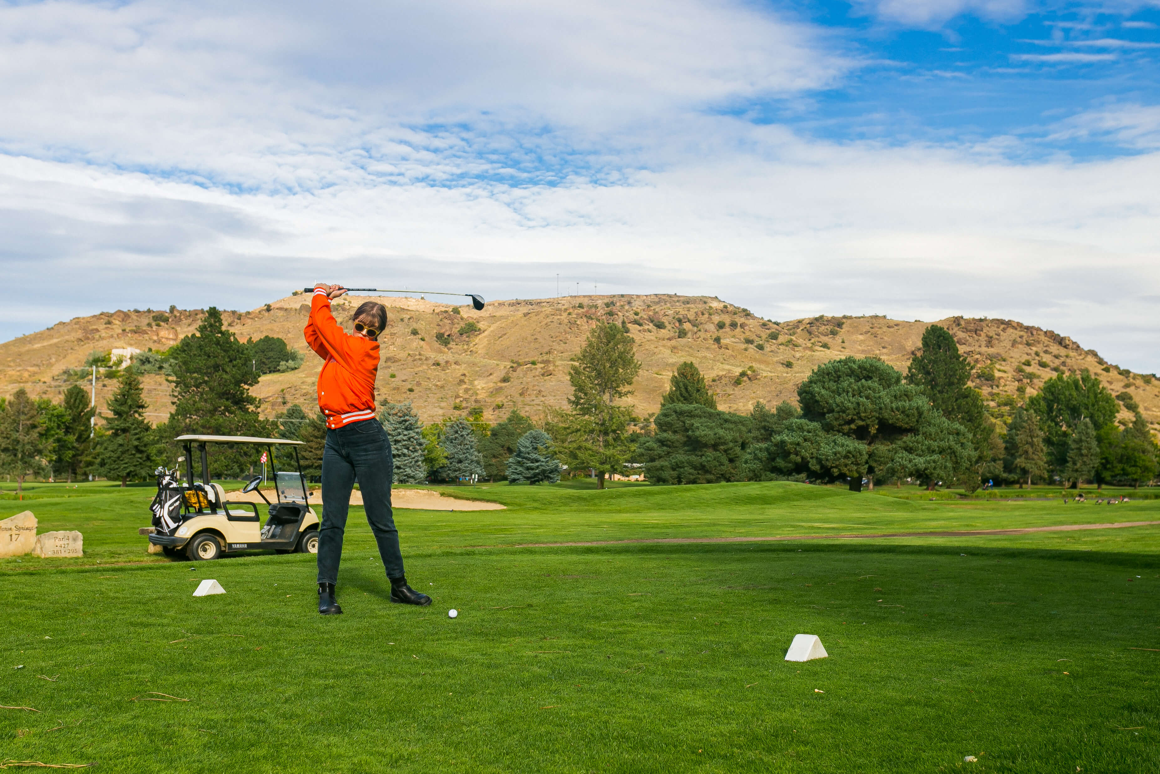 Woman in orange jacket swinging golf club at Warm Springs Golf Course with blue skies and green grass.