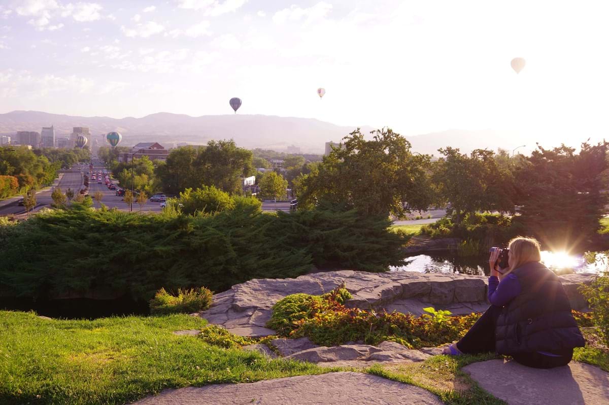 man and woman sitting on ledge watching the sun rise and hot air balloons in the sky