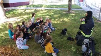 Group of about 20 school children sit in grass while Boise Firefighter puts on full suit