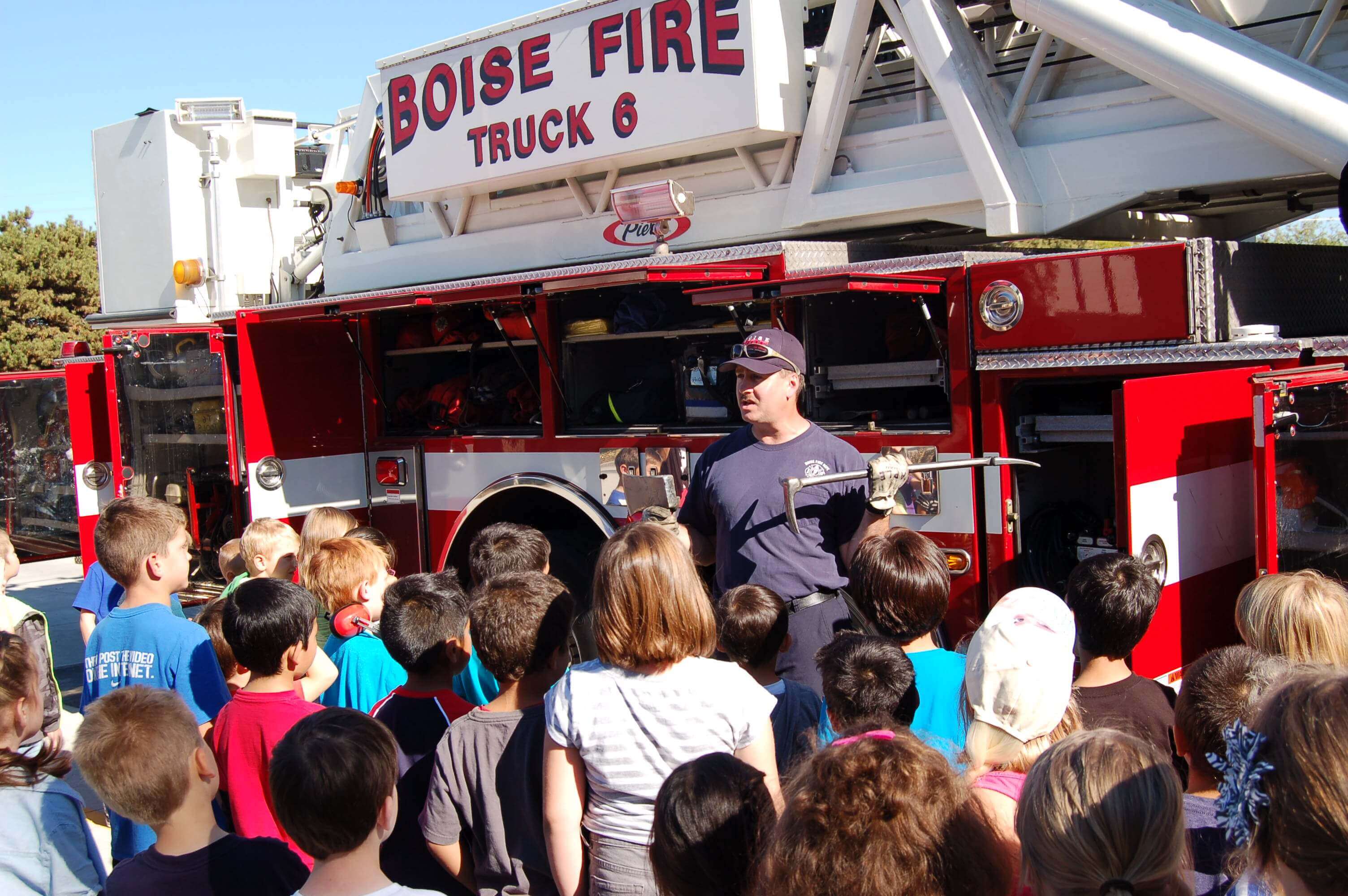 A firefighter stands in front of a fire engine and gives a presentation to a group of children.