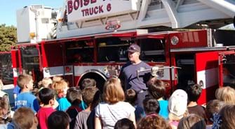 Firefighter holds tools with truck in background and kids crowd around to look