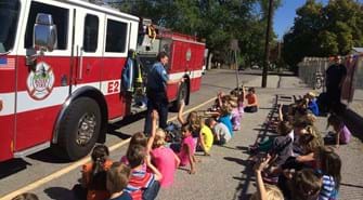 Kids sit on ground in front of fire truck as a firefighter walks in front of them answering question