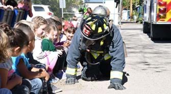 Firefighter crawls on ground in full gear, including mask, so children can see him up close