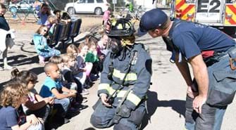 Firefighter in full gear kneels in front of students. Another firefighter looks on.