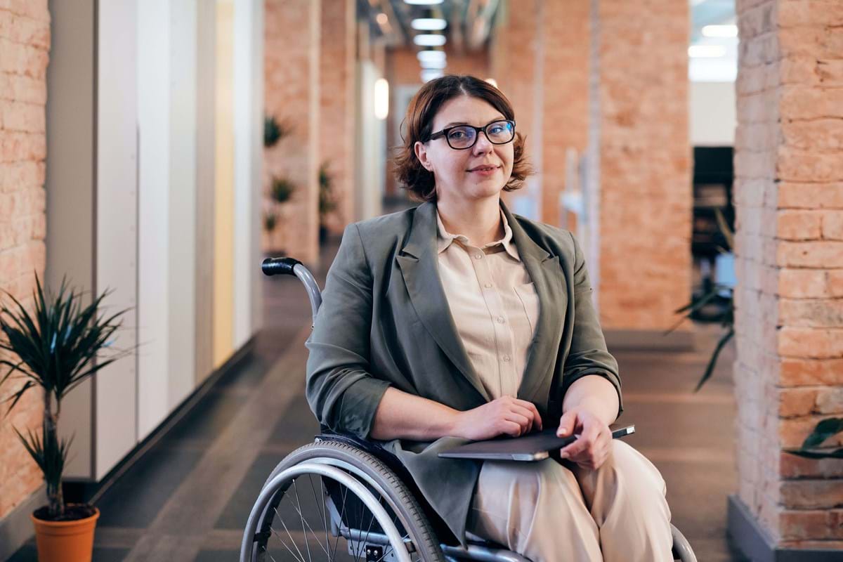 Woman with dark hair and glasses sitting in wheelchair inside a brick building, smiling at camera.