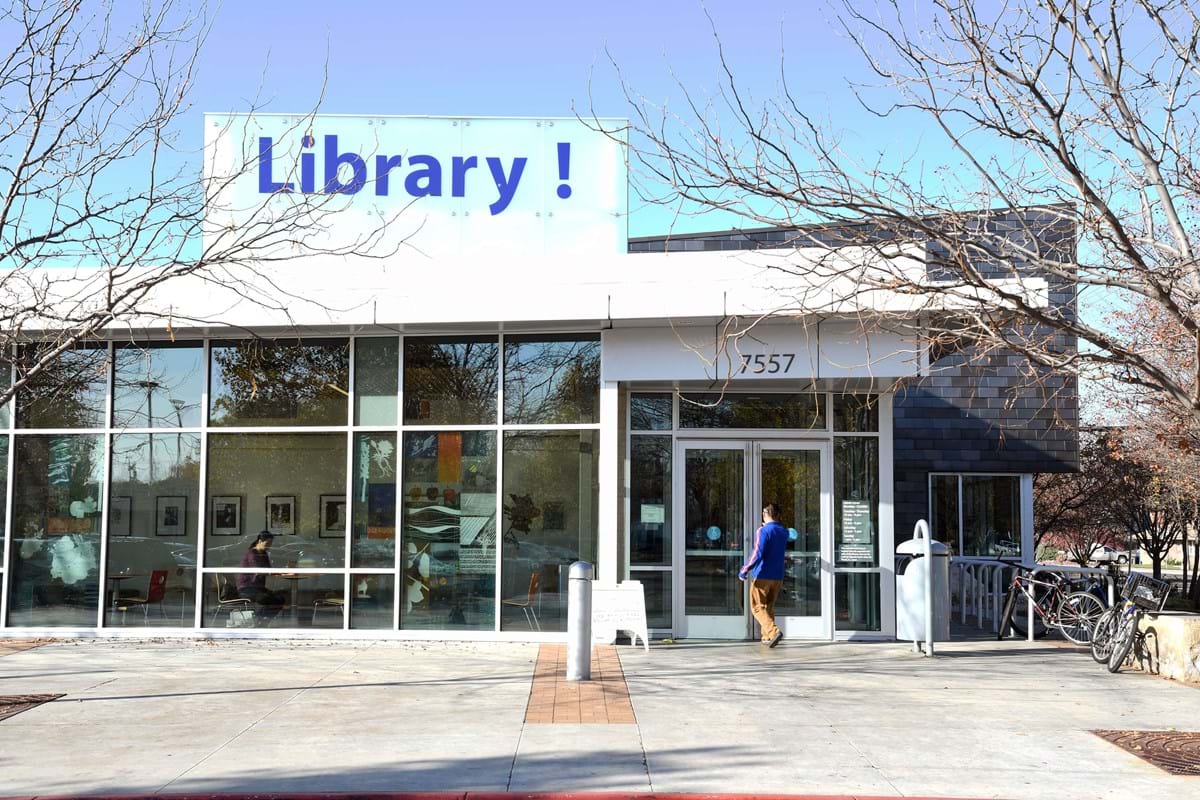 Exterior of Cole and Ustick Library with blue Library! sign on top and clear glass windows. Trees surrounding the building without leaves and man walking to front door.