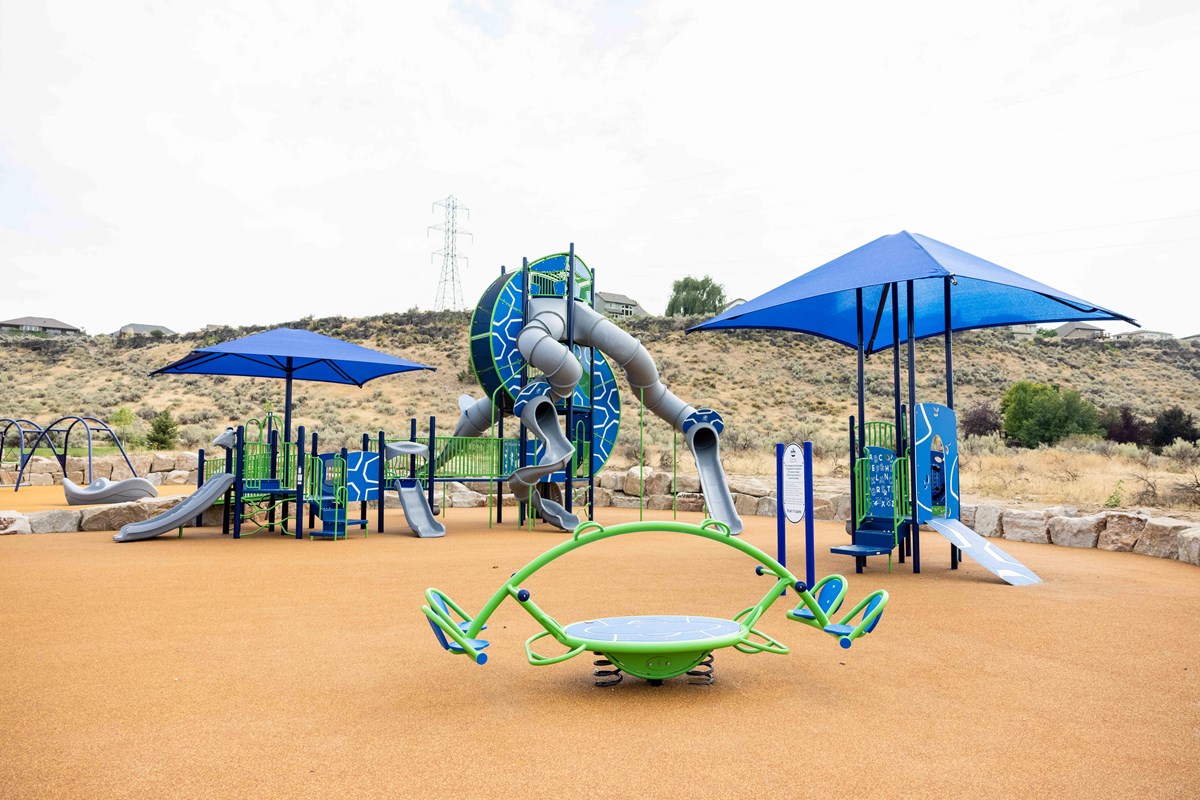 First, Bowler Park depicting tan fall material and blue and green ADA Accessible play equipment.