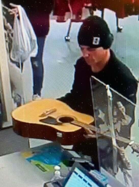 man wearing black jacket and black beanie hat standing at shop counter holding a guitar