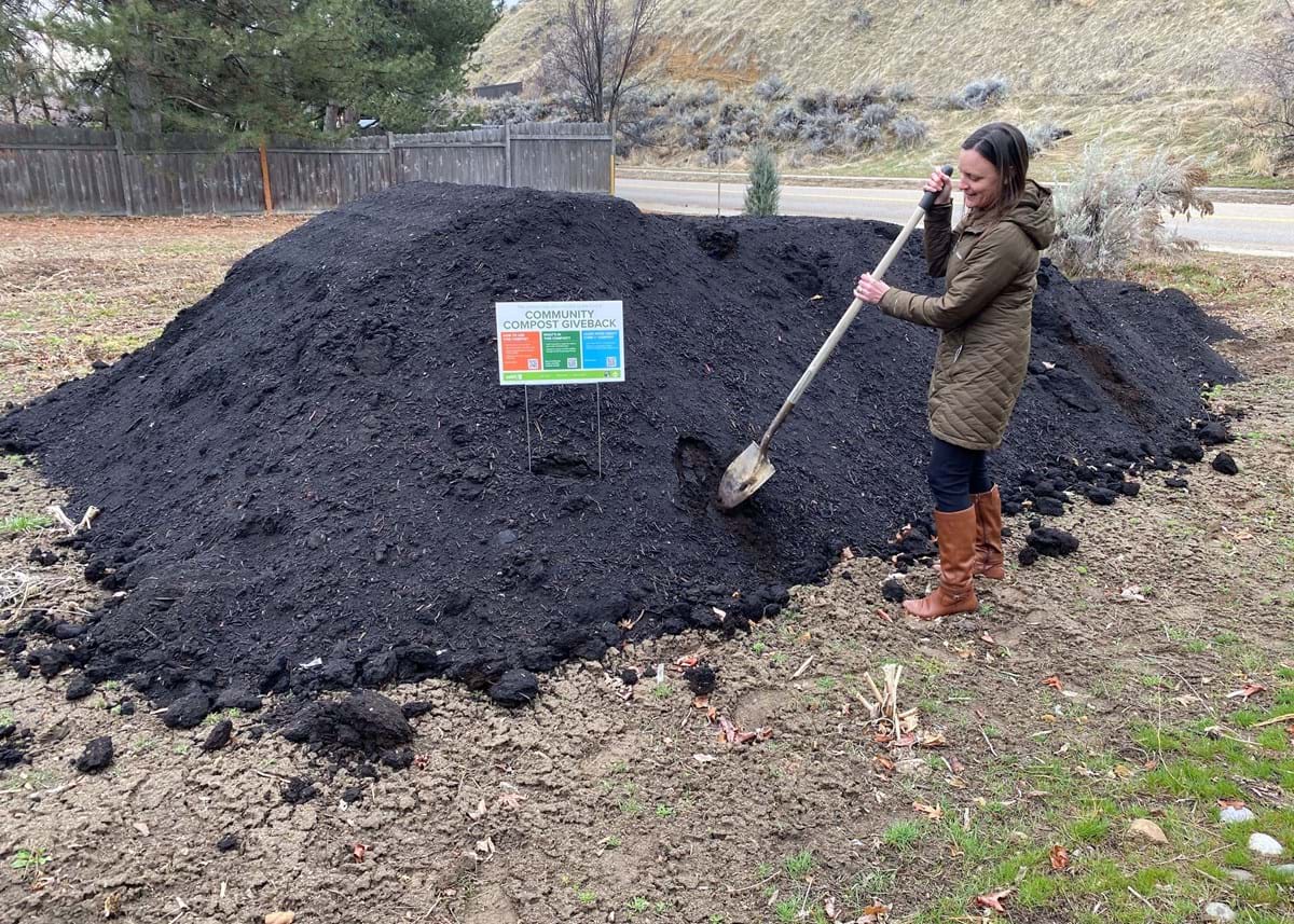 Person standing next to a large pile of compost, holding a shovel.