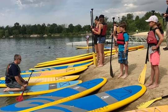 Group of kids learning to paddle board standing on the edge of a pond.