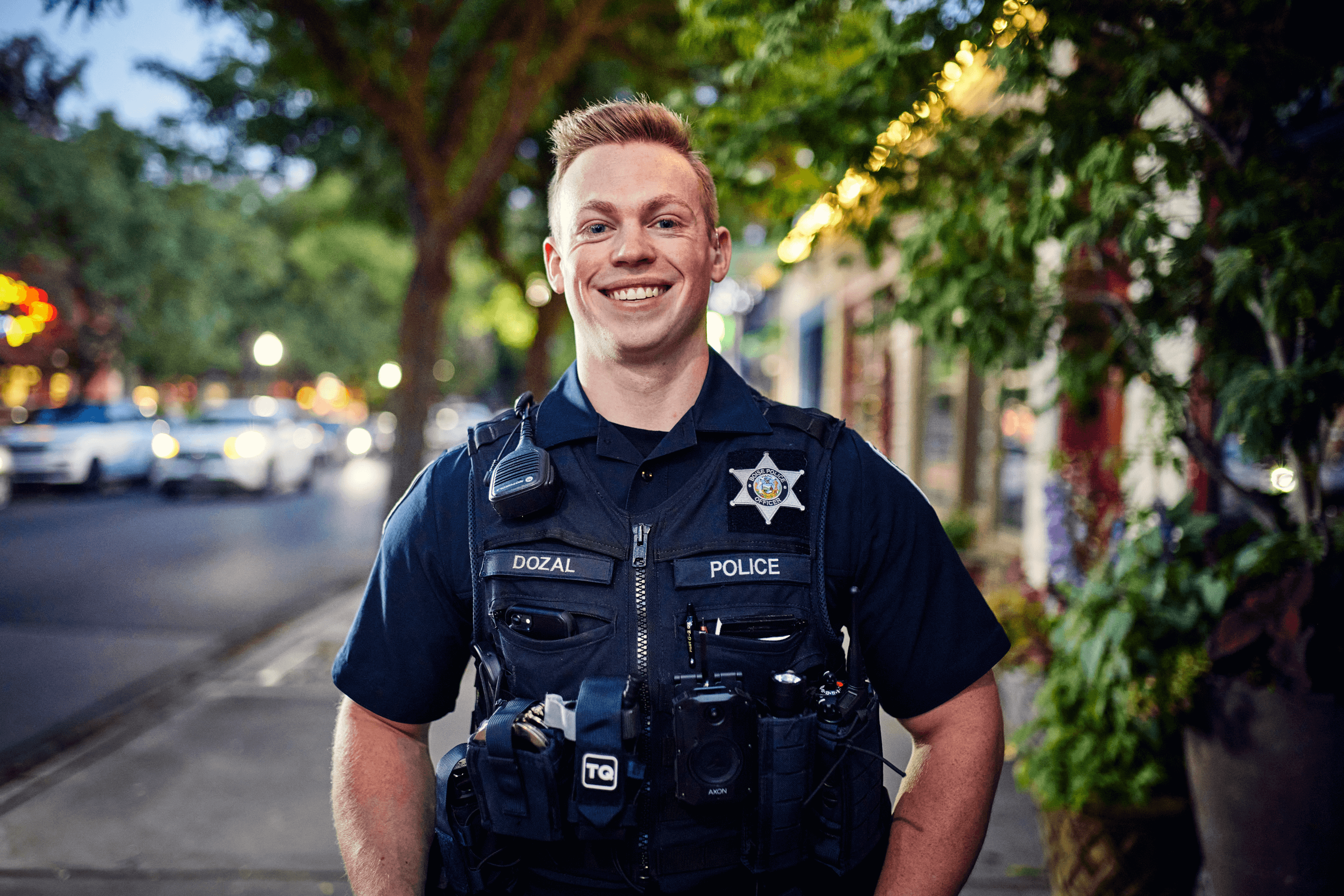 Officer smiling at the camera with green trees behind him standing on a sidewalk in the evening.