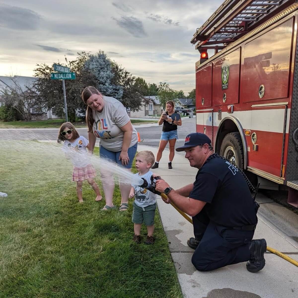 Firefighter kneeling down next to a fire truck showing kids the fire hose.