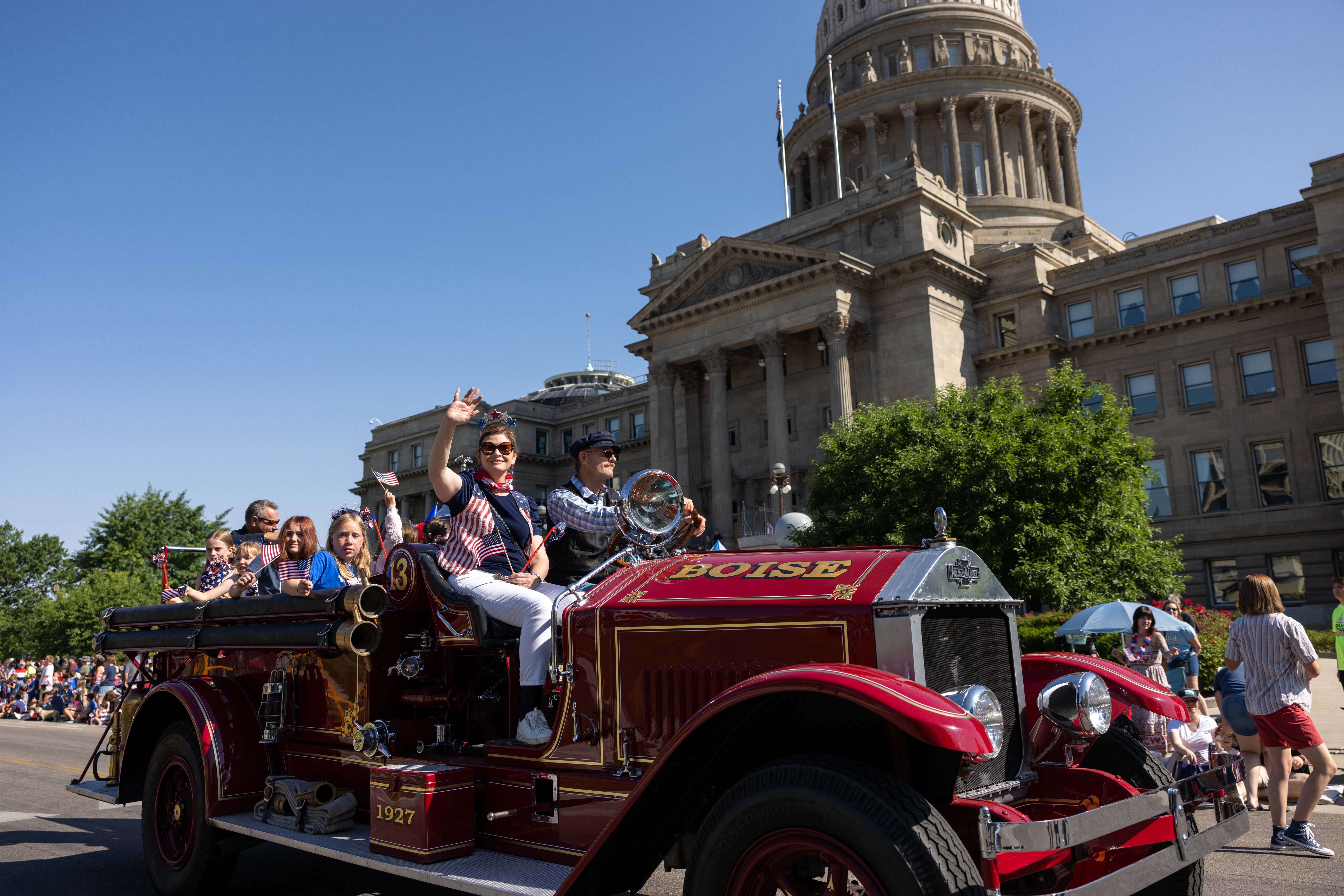 People riding an old fire engine during the 4th of July parade with the Capitol in the background.