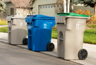 A trash, recycling and compost bin sit on a curb on a neighborhood street during the day.