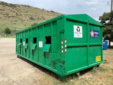 A large green drop off bin at a drop-off location on a dirt road during the day.