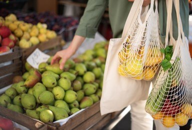 A person picks up a pare at a grocery store while holding reusable bags full of assorted fruits.