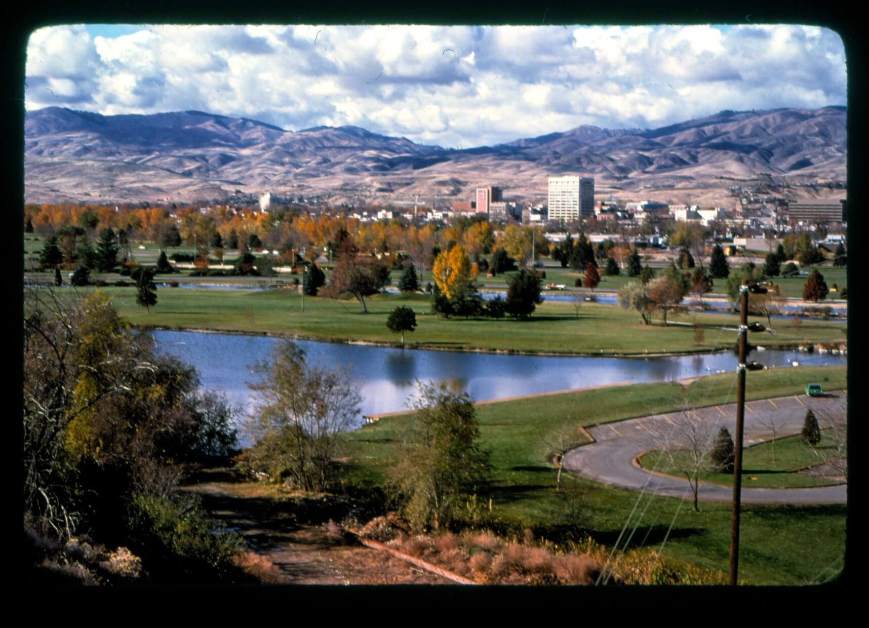 View of Boise looking out over a park space with pond, green grass, trees, city skyline and foothills