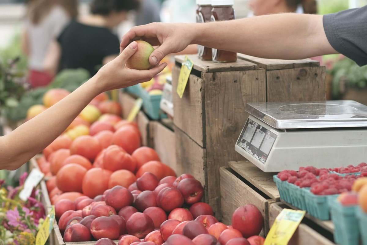 Close up photo of person handing a peach to someone else over a shelf of other fruits at a farmer's market